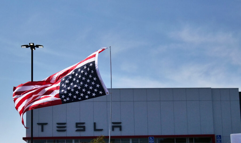 This photo shows a large American flag waving prominently in front of a Tesla building. The building has the recognizable Tesla logo on it, and the sky is clear and blue in the background. There are also visible handicap parking signs and a tall light post