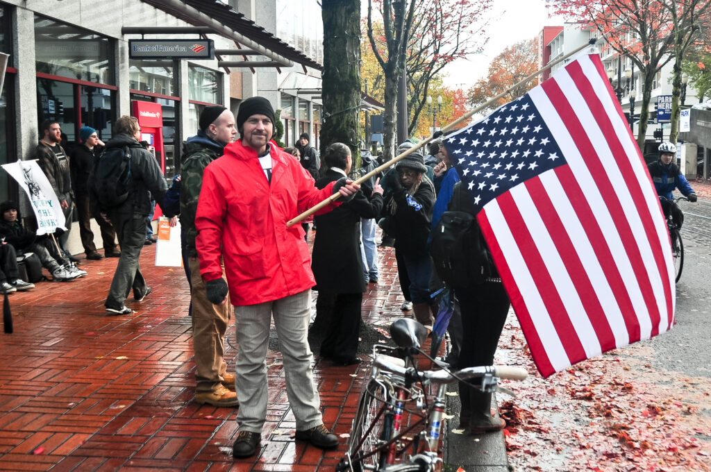 A man in a red jacket holding an American flag during a street protest in front of a Bank of America building. Several people are gathered around, some with signs, on a wet sidewalk covered in autumn leaves