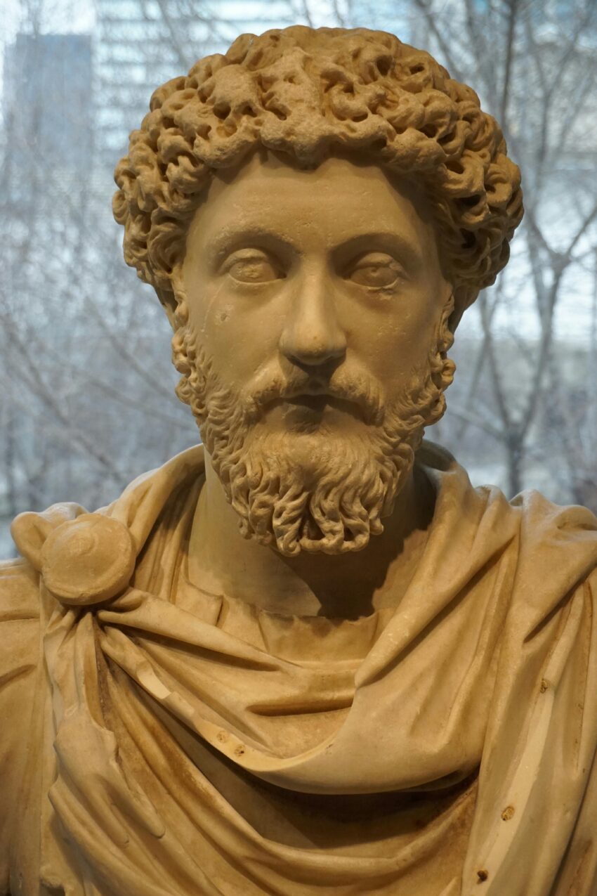A marble bust of a bearded man with curly hair, dressed in a classical Roman toga. The sculpture is finely detailed, capturing lifelike facial features and textured hair. The background shows blurred trees and a modern building, suggesting the statue is displayed indoors near a window. A marble bust of a bearded man with curly hair, dressed in a classical Roman toga. The sculpture is finely detailed, capturing lifelike facial features and textured hair. The background shows blurred trees and a modern building, suggesting the statue is displayed indoors near a window. A marble bust of a bearded man with curly hair, dressed in a classical Roman toga. The sculpture is finely detailed, capturing lifelike facial features and textured hair. The background shows blurred trees and a modern building, suggesting the statue is displayed indoors near a window.